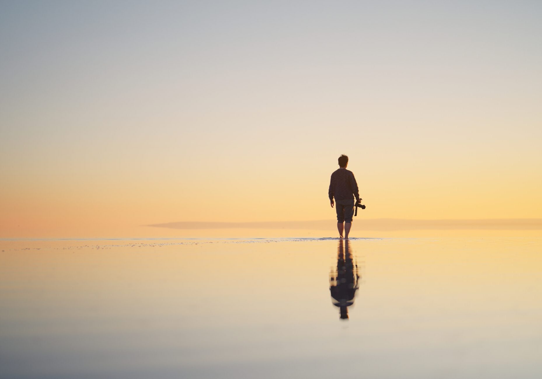 A rear view of a man solo traveler holding his camera and walking on the shallow surface of Salt Lake, Tuz Golu during sunset in Turkiye.