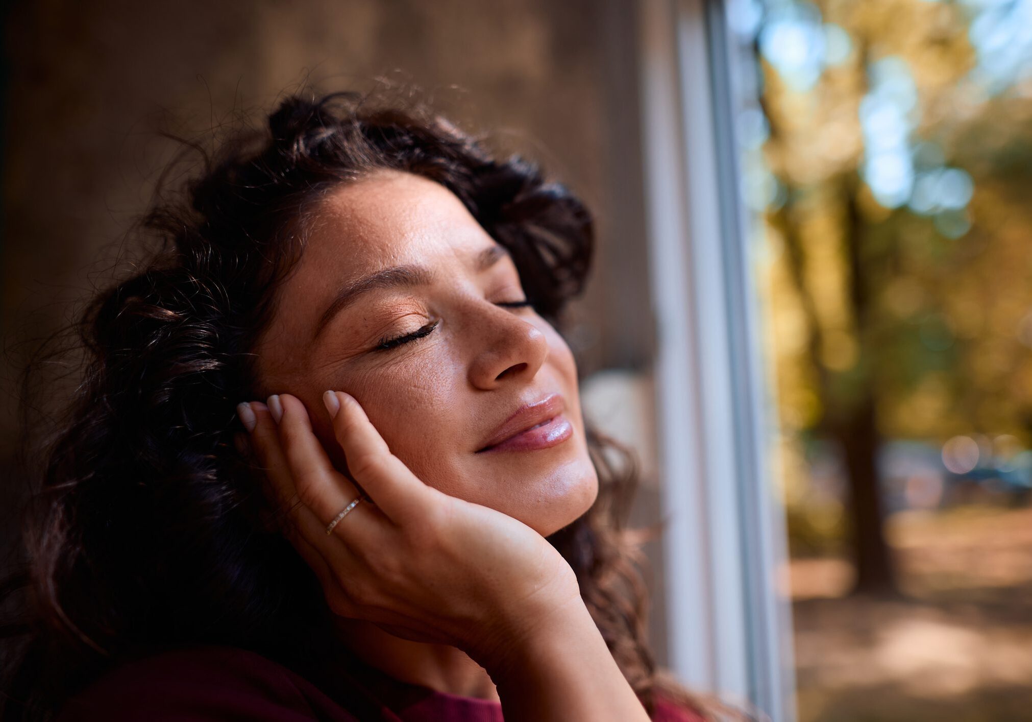 Mid adult woman with curly hair and closed eyes gently holds her face, finding tranquility and enjoying a peaceful moment by the window with a blurred autumnal view in the background